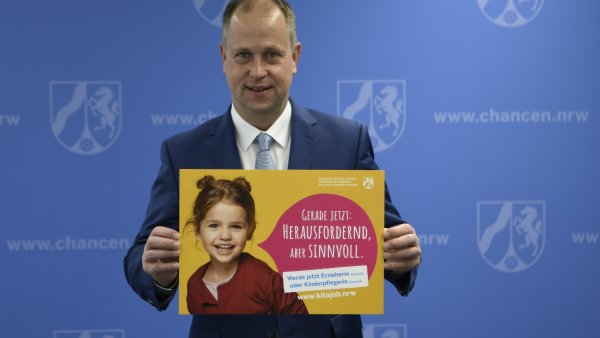 Ein Mann im Anzug steht vor einer blauen Wand und hält ein gelbes Plakat hoch, auf dem ein Kind zu sehen ist