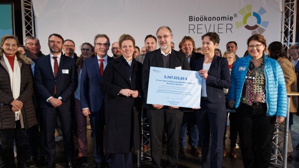 Vor einer Leinwand mit dem Bioökonomie Revier Logo stehen viele Menschen. Die vorderen beiden halten ein Schild hoch.