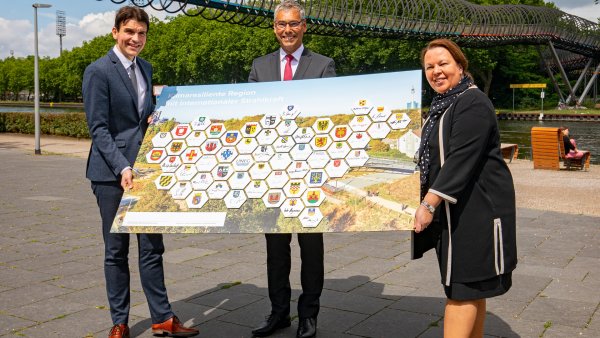 Zwei Männer und eine Frau stehen an einer Uferpromenade und halten ein Schild mit Städte-Wappen hoch