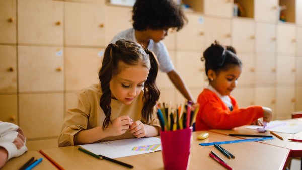 Drei Kinder sitzen an einem Holztisch und beschäftigen sich mit Zeichnen und Malen; im Vordergrund zeichnet ein Mädchen mit Zöpfen, während zwei weitere Kinder im Hintergrund kreativ arbeiten. Auf dem Tisch stehen ein roter Becher mit Buntstiften sowie verschiedene Stifte, Lineale und Zeichenmaterialien.