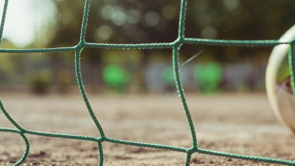 Nahaufnahme eines grünen Fußballnetzes, das im Vordergrund scharf zu sehen ist. Im unscharfen Hintergrund erkennt man einen Ball am rechten Bildrand und eine Spielfläche im Freien.