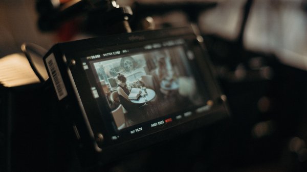 Eine professionelle Filmkamera zeigt auf ihrem Monitor eine Filmszene, in der eine Frau an einem Tisch sitzt und in einem Buch liest. Die warme, stimmungsvolle Beleuchtung und der unscharfe Hintergrund vermitteln einen cineastischen Blick hinter die Kulissen einer Filmproduktion.