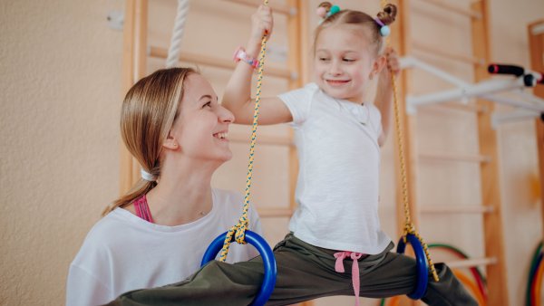 Ein Kind mit weißem T-Shirt und dunkler langer Hose trainiert an Gymnastikringen, während eine Lehrerin es aufmerksam begleitet und motivierend anlächelt.