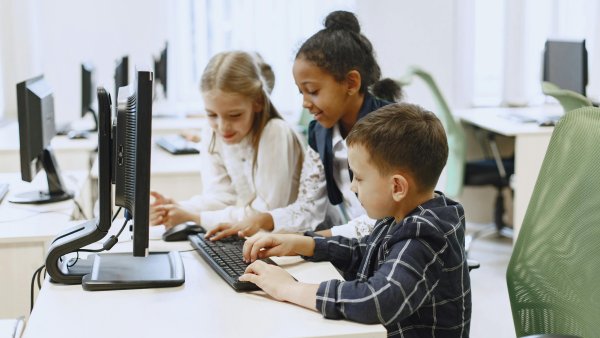 Zwei Mädchen und ein Junge im Grundschulalter sitzen in einem Computerraum gemeinsam vor einem Bildschirm, der Junge tippt etwas auf der Tastatur