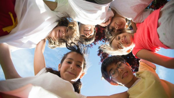 Eine Gruppe von Kindern steht im Kreis und blickt lächelnd nach unten in die Kamera. Die Aufnahme ist von unten gemacht, sodass der Himmel und etwas Baumlaub im Hintergrund zu sehen sind. Die Kinder wirken fröhlich und halten vermutlich zusammen oder lehnen sich zueinander.