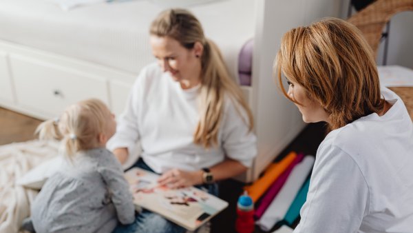 Eine Frau, wie eine weitere Frau mit einem kleinen Kind gemeinsam ein Bilderbuch anschaut. Die drei sitzen in einer hellen, wohnlichen Umgebung und wirken aufmerksam miteinander im Gespräch.