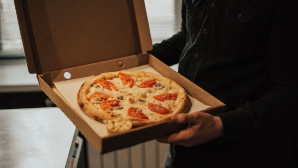 Ein Mensch mit schwarzer Kleidung (Kopf ist nicht sichtbar) hält eine Packung eines Pizza-Lieferdienstes in der Hand, die offen ist und eine Pizza mit Tomaten zeigt. Hintergrund ist eine Heizung und ein kleiner Kühlschrank erkennbar.