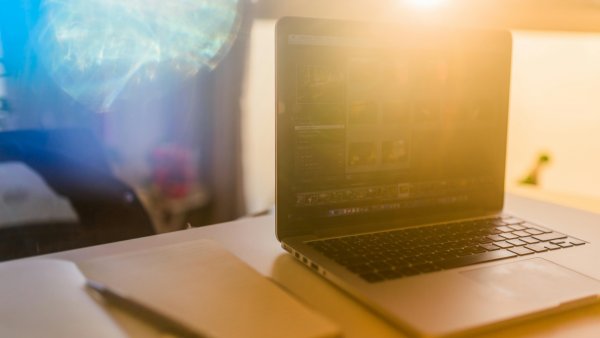 auf einem hellen Schreibtisch liegen ein Notizbuch mit einem Stift links und recht ein aufgeklappter Laptop, auf dem verschwommen ein dunkler Bildschirm mit Zahlen zu erkennen ist. Im Hintergrund scheint durch das Fenster die Sonne und eine Spiegelung der Sonne verdeckt den restlichen Hintergrund.