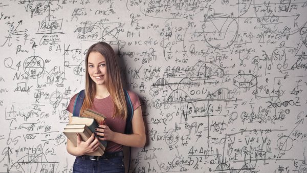 Eine junge Frau steht in Jeans-Latzhose und einem rötlichem T-Shirt mit mehreren Büchern in den Armen vor einer Tafel voller handschriftlicher mathematischer Formeln und Diagramme.