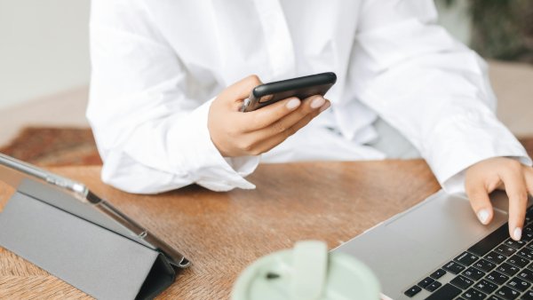 Person in weißer Bluse sitzt am Schreibtisch und nutzt gleichzeitig Smartphone und Laptop. Auf dem Tisch stehen außerdem ein Tablet in Schutzhülle und ein Becher – modernes Arbeiten im Homeoffice oder Büro.