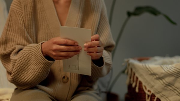 Person in beigem Strickpullover hält einen Brief in den Händen und sitzt auf einem Sofa. Im Hintergrund steht eine Pflanze, rechts ist ein Korbstuhl mit einer Decke zu sehen.