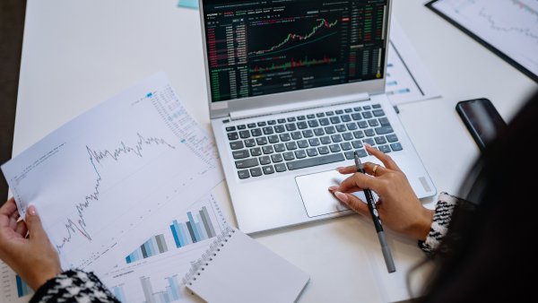 Person analysiert Börsencharts auf einem Laptop und vergleicht diese mit ausgedruckten Diagrammen. Die Szene zeigt die Auswertung von Finanz- und Marktdaten.