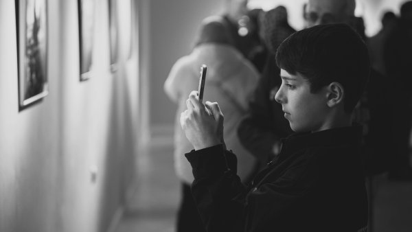 Ein Junge steht in einer Galerie und fotografiert konzentriert ein Kunstwerk mit seinem Smartphone. Im Hintergrund sind weitere, unscharfe Besucher zu sehen, die sich ebenfalls die Ausstellung ansehen. Die Szene ist in Schwarz-Weiß gehalten und wirkt ruhig und aufmerksam.