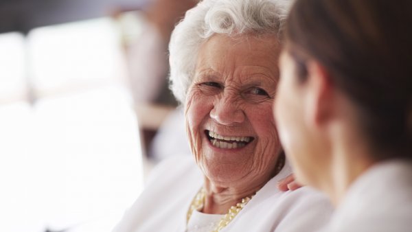 Das Bild zeigt eine lächelnde alte Frau im Gespräch mit einer anderen Person.