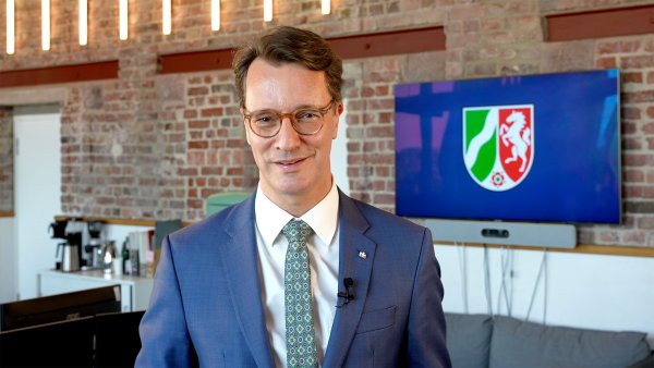 MP Wüst steht in einem Großraumbüro mit einer NRW-Flagge im Hintergrund und schaut in die Kamera