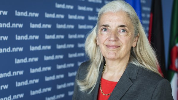 Ministerin Isabel Pfeiffer-Poensgen lächelnd vor EU-, Deutschland- und NRW-FLaggen