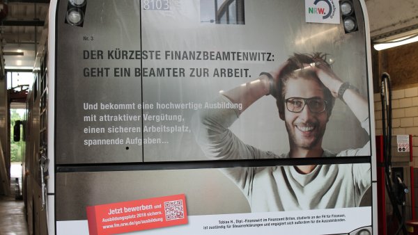 Ein Plakat auf der gesamten Rückseite eines Buses aufgeklebt. Ein Mann mit Brille rauft sich die Haare. Links daneben der Witz: "Geht ein Beamter zur Arbeit..."