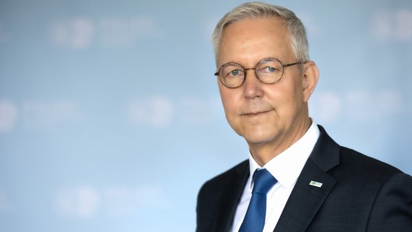 Oberkörper eines Mannes mit grauen Haaren, runder Brille und einem schwarzen Anzug mit dunkelblauer Krawatte und weißem Hemd. Im Hintergrund ist unscharf eine Logowand der Polizeibehörde Dortmund zu erkennen, weiße Schrift auf hellblauem Untergrund.