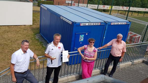 Staatssekretärin MIlz rosa und pink gekleidet steht mit MItgliedern vom TuS Oberpleis 1905 e.V. an einem hellgrauen Metallgitterzaun gelehnt.