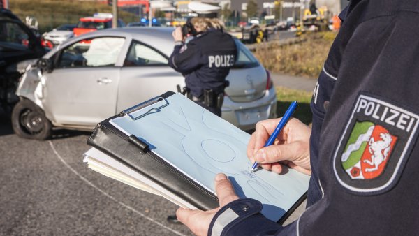 Ein Polizist hält ein Klemmbrett in der Hand, auf dem er einen Unfallhergang aufzeichnet. Im Hintergrund fotografiert eine Polizistin zwei Autos, die zusammengestoßen sind, dahinter ist ein Stau zusehen.