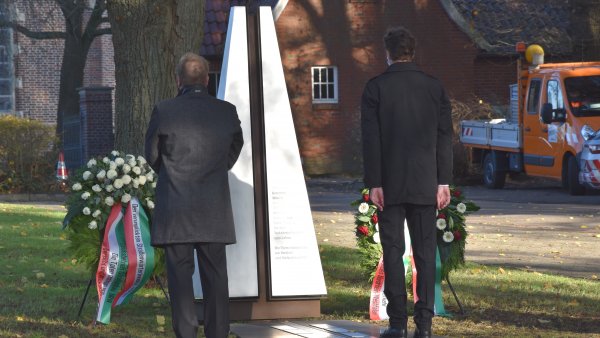 Zwei Männer stehen vor einem Gedenkstein, der mit zwei Blumenkränzen geschmückt ist