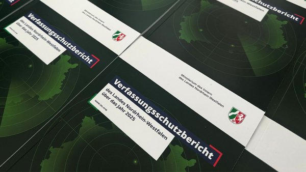 Mehrere Exemplare eines gedruckten Berichts liegen nebeneinander auf einem Tisch. Auf den Titelseiten steht „Verfassungsschutzbericht des Landes Nordrhein-Westfalen über das Jahr 2025“ sowie „Ministerium des Innern des Landes Nordrhein-Westfalen“ mit Landeswappen. Das Design zeigt eine grüne Karte mit kreisförmigen Markierungen auf dunklem Hintergrund.