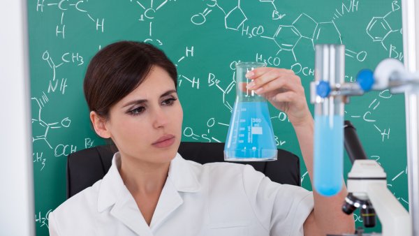 Eine dunkelhaarige Frau im weißen Kittel hält einen Glaskolben mit hellblauer Flüssigkeit in der linken Hand und schaut es prüfend an. Im Hintergrund eine grüne Tafel mit chemischen Formeln in weißer Kreideschrift.