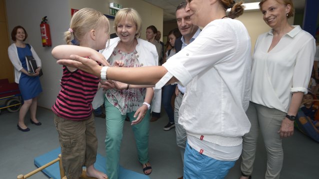 Ein Kind geht über einen Barren und wird von der Erzieherin gestützt. Im Hintergrund lacht Ministerpräsidentin Hannelore Kraft.