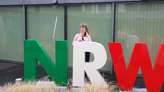 Staatssekretärin Andrea Milz steht vor einer Fensterfront, vor ihr die drei Buschtaben N R W in grün, weiß und rot.