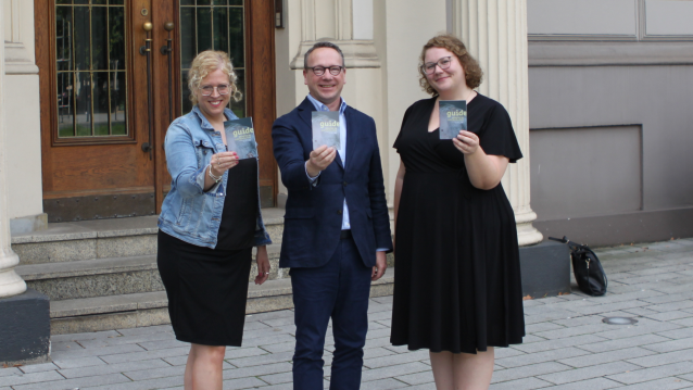Zwei Frauen und ein Mann stehen vor einer Eingangstür und halten ein Ratgeberheft hoch