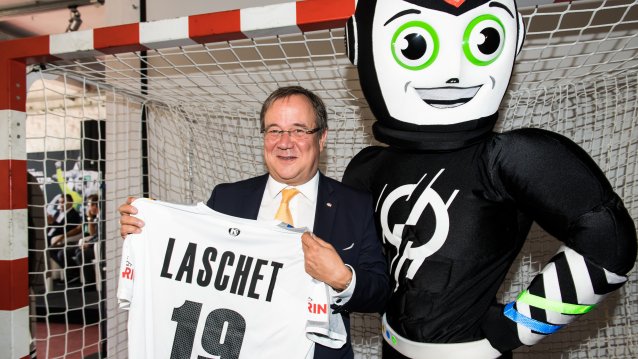 Ministerpräsident Laschet steht mit Maskottchen im Handballtor, er hält ein weißes Handballer-T-Shirt mit seinem Namen "Laschet" und der Nummer 19 ausgebreitet in seinen Händen.