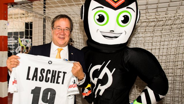 Ministerpräsident Laschet steht mit Maskottchen im Handballtor, er hält ein weißes Handballer-T-Shirt mit seinem Namen "Laschet" und der Nummer 19 ausgebreitet in seinen Händen.