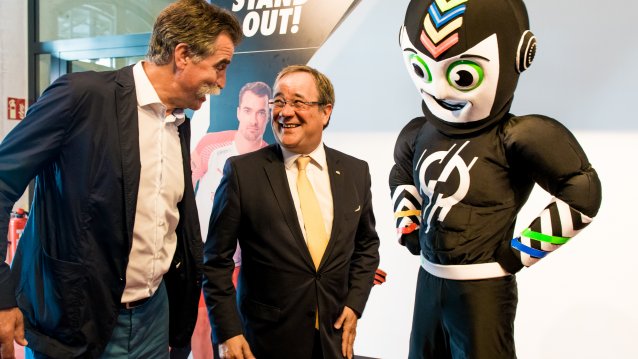 Ministerpräsident Laschet im Gespräch mit einem Mann, rechts neben ihm das Maskottchen.
