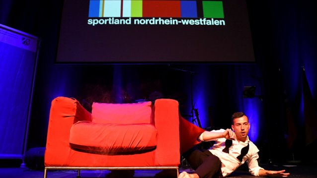 Showakt - Auf der Bühne steht ein großer roter Sessel, rechts daneben liegt ein Mann auf einem Arm abgestüt, den anderen Arm angewinkelt. Er hat ein weißes Oberteil und eine schwarze Hose an.