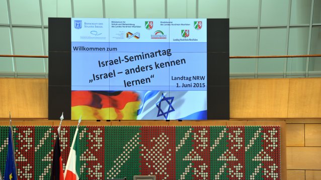 Über dem rednerpult hängt ein Monitor mit dem Titel der Veranstaltung "Israel-Seminartag. Isreal anders kennenlernen"