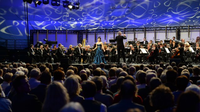 Eine Opernsängerin steht neben dem Dirigenten, im Hintergrund das Orchester, im Vordergrund Köpfe des Publikums