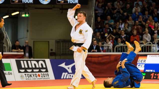 Nach einem erfolgreichen Wurf streckt der Judoka im weißen Anzug seinen rechten Arm mit geballter Faust nach oben, während der Judoka im blauen Anzug auf den Boden liegt.