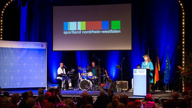 Staatssekretärin Andrea Milz am Rednerpult rechtsseitg auf einer Bühne. Links sitzt eine Musikband. Vor der Bühne sitzt das Publikum.