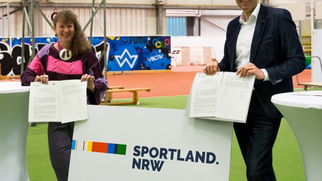 Links im Bild steht Staatssekretärin Andrea Milz, sie  hat eine pink-schwarze Sportjacke an. Rechts im Bild steht ein Mann im schwarzen Anzug und weißem Hemd. Zwischen ihnen steht unten auf dem Boden ein  weißer Austeller mit dem "Sportland NRW"-Logo. Sie befindne sich in einer Indoor-Sporthalle.