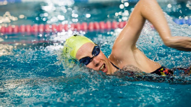 Eine Wettkampfschwimmerin im schwarzen Badeanzug mit schwarzer Schwimmbrille und  gelber Bademütze krault durchs Wasser.