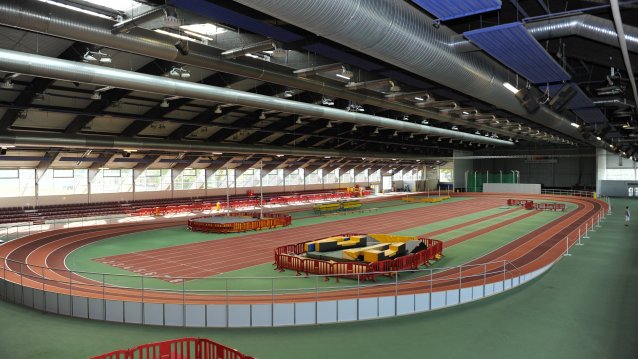 Leichtahtletiksporthalle von  innen mit einer umlaufenden, roten Tartanbahn und einer geraden, roten Tartanbahn in der Mitte. Mit weiteren Sportanlagen wie einer Hochsprunganlage. Das Dach ist geschlossen.