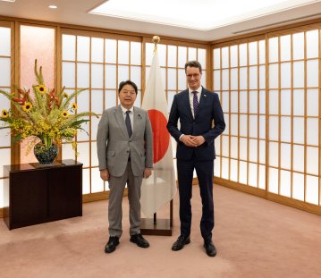 Zwei Männer in Anzügen stehen in einem japanischen Raum mit Japan-Flagge und einer Kommode und Blumen im Hintergrund