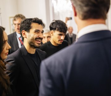 Fußballer İlkay Gündoğan trägt einen dunklen Anzug und lacht zwischen anderen Personen in Business-Kleidung.