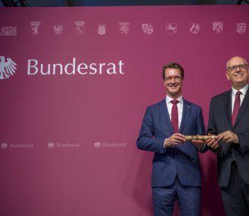 Zwei Männer in Anzügen halten gemeinsam einen Holzstab fest.