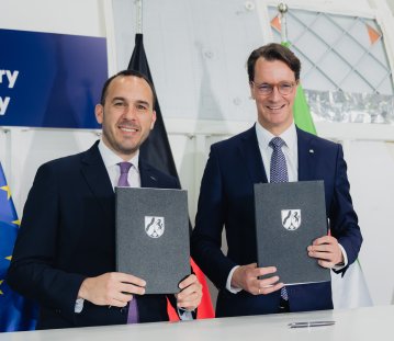 Ministerpräsident Wüst und ein Mann halten das unterzeichnete Papier NRW-Mappen in die Kamera