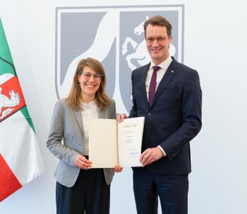 Rechts neben der NRW-Fahne steht Verena Schäffer mit langen blonden Haaren und Brille im grauen Sakko und weißem Hemd lächelnd neben MP Wüst im schwarzen Anzug mit roter Krawatte