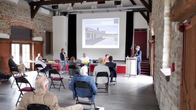 In einem Saal wird ein Vortrag mit einer Päsentation auf einer Leinwand gehalten. Der Redner steht rechts vorne an einem Rednerpult.