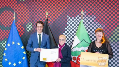 Ein Mann und zwei Frauen stehen vor der Europa-, Deutschland- und NRW-Flagge