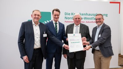 MP Wüst und Minister Lauman posieren mit Förderbescheidempfängern und eine Urkunde für ein Foto