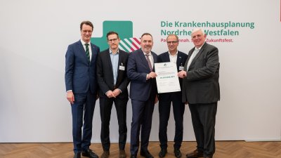 MP Wüst und Minister Lauman posieren mit Förderbescheidempfängern und eine Urkunde für ein Foto